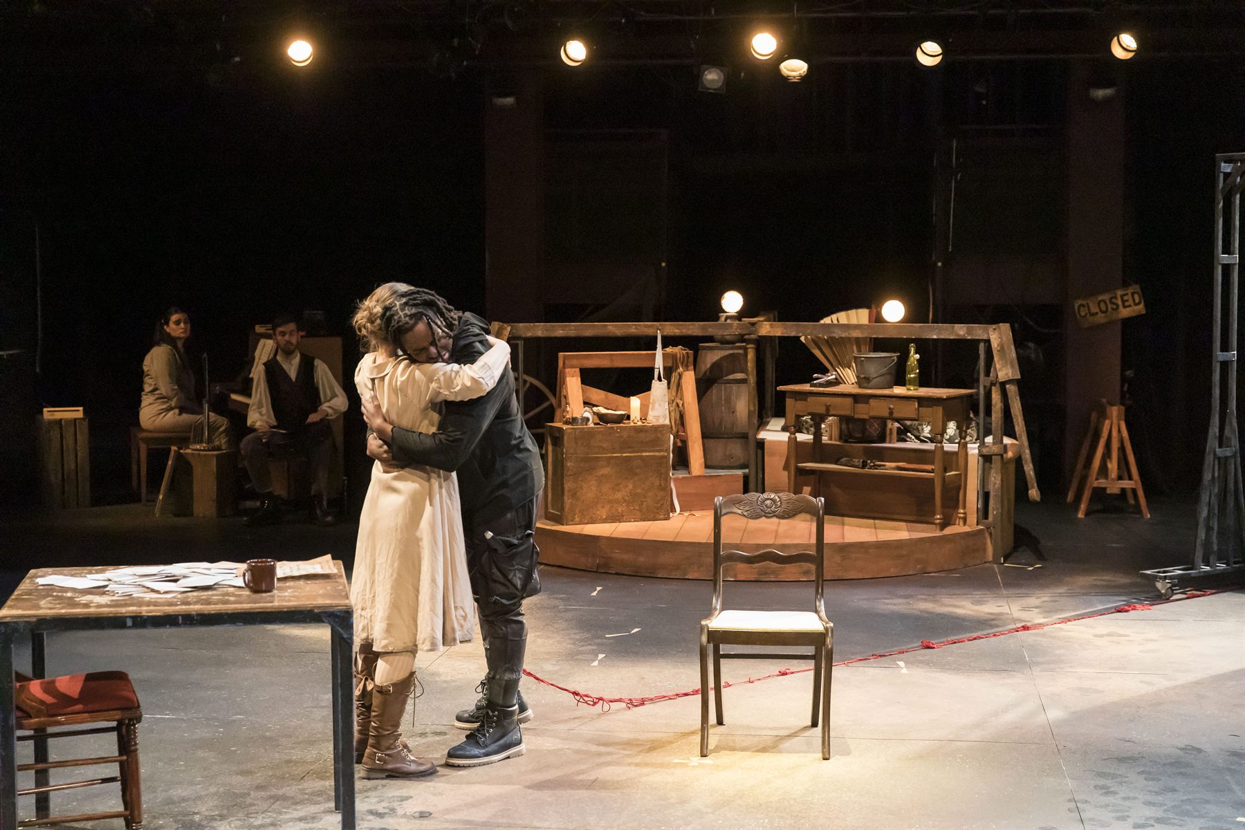 Two figures embrace at center stage under practical bulbs